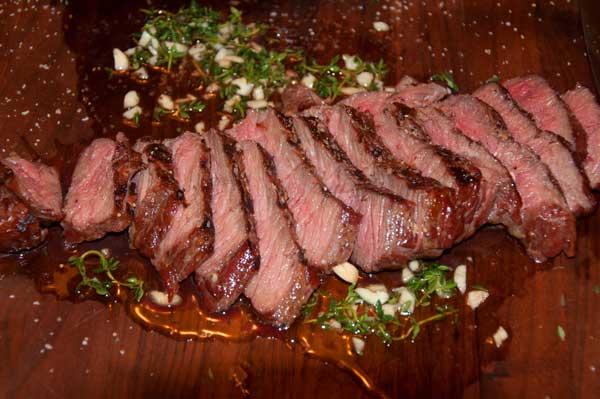 Grilled, sliced steak in oil and chopped green herbs on a wood cutting board