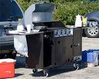 Large black gas grill with shiny steel lid up in a parking lot surrounded by cars. Food and cooking utensils are on the shelves attached to both sides. A red cooler sites on the ground to the left of the grill.