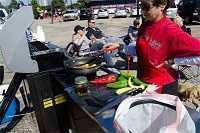 Woman in a red shirt cooking on a large gas grill in a parking lot surrounded by people and cars.