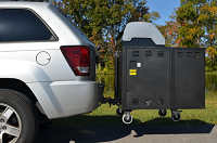 A large black box with wheels raised off the pavement and hitched to the back of a white car.