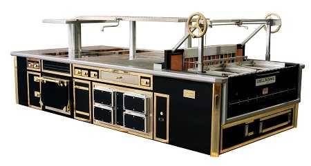 Very large contraption made of black metal, ceramic, brass and stainless steel. It looks like an enormous kitchen island with drawers, doors and built in ovens. A large bar connected to a crank wheels is suspended above.