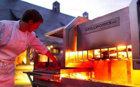 A man in a white cook's apron tending a roaring wood fire in a very large shiny steel enclosure.