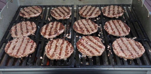 Hamburgers cooking on a gas grill with pronounced brown sear marks on the meat surface.