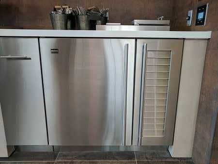 Shiny stainless steel doors and drawers under a kitchen counter with small buckets filled with napkins on top. Alight switch is on the wall to the right.