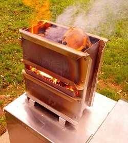 Small metal box filled with burning wood on top of a larger metal box, green grass in the background.