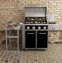 Combination black and shiny steel gas grill placed in front of a brick wall. The lid is up showing food cooking. A small table on the left side also has food on it.