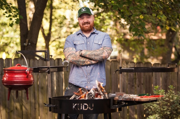 A bearded man with a red hat stands behind a black metal bowl on legs filled with burning wood.