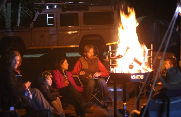 People stiing around a blazing fire bowl at night. An auto mobile is in the background.