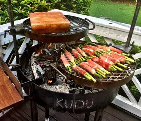 Black metal fire bowl on an outdoor patio. A wood fire is inside the fire bowl. Two round cooking grates with food are positioned above the fire.