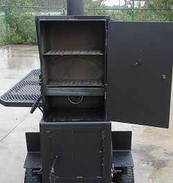 Large black cabinet with door open showing two cooking racks. A chimney comes out of the top It sits on pavement with green bushes in the background.