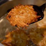 A crispy potato latke lifted from the oil on a slotted spoon