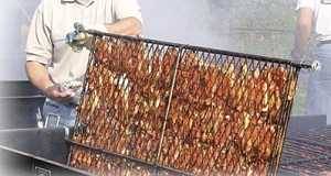 A man flips a double BBQ cooking grate that has a load of chicken parts sandwiched between the two grates. He is cooking the chicken over a black grill.