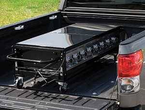 Large 8 burner gas grill with legs folded up to compress the entire system into a flat rectangle and placed in the back of a pick up truck.