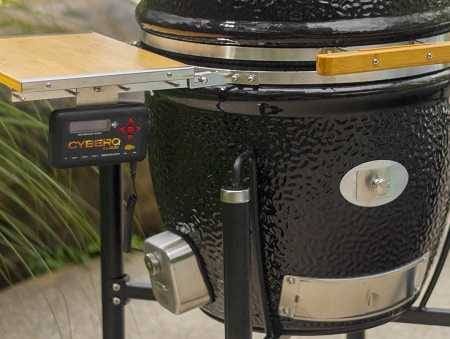 Black kamado on right with a wood shelf attached on the left. An black box labeled "Cyber Q" hangs from the shelf. There are green shrubs in the background.