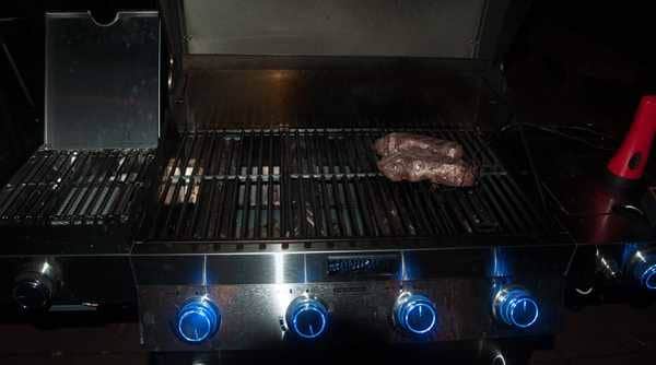 A gas grill at night. The lid is up showing two steaks cooking on the right side. The gas burner control knobs have blue lights.