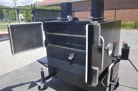 Big cabinet smoker in parking lot with wire fence, brick building and trees in the background. Two doors are open showing the cabinet interior with two slide out racks.