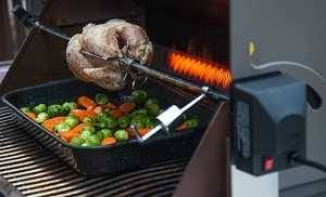 A gas grill with the lid up. A rotisserie system is cooking a whole chicken over a tray of vegetables. A glowing burner is behind the chicken.