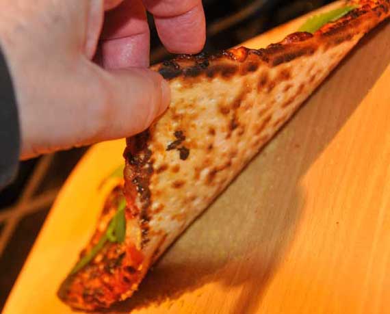 A hand holds up a piece of pizza to show the bottom crust.