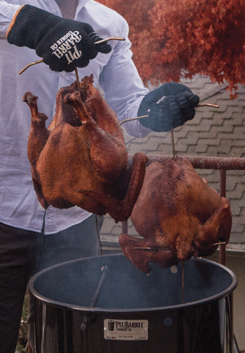 A man wearing black golves is pulling two smoked turkeys on steel hooks from a smoking barrel. Colorful autumn trees are in the background.