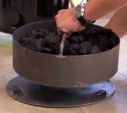 A man holds a metal basket by a handle. The basket is filled with charcoal. There is a metal disc under the basket.