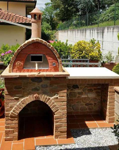 Large outdoor brick fireplace witha wood storage area on the right and a pizza oven with chimney on top. A house with clay tile roof and green trees are in the background.