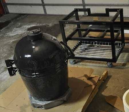 Large black kamado on wood shipping pallet on left and large black cart frame on right. Inside a garage.