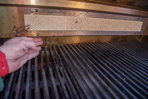 A bbq grill surface with a metal, overhanging part above. A hand reaches in from the left under the overhanging part.