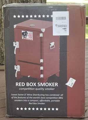 Large colorful carboard box on an outdoor deck. A photo of a red cabinet smoker is on the box. "Red Box Smoker" is printed under the photo.