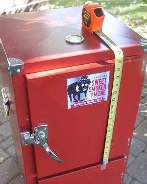 Red upright cabinet on an outdoor patio. There is a metal plate on the front with a picture of an animal reading"Swet Smoker Mine". Measuring tape is pulled from the bottom to top. There is a shiny steel latch on the right.