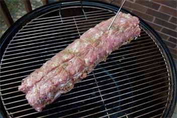 Round charcoal grill on an outdoor deck. The lid is off showing a long, seasoned pork loin on the cooking grate.