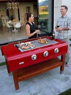 A man ad woman on an elegant patio drinking wine. A dining room with chandelier is in the back ground, They are standing arouns a large red gas grill with no lid. The shiny steel cook surface has crab legs and shish kabob.