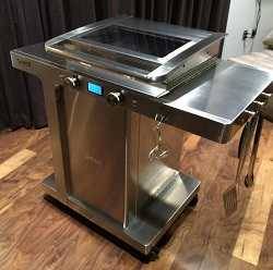 Shiny steel gas grill on a wood floor with a black curtain in the background. The grill lid is flat and has a window in it.