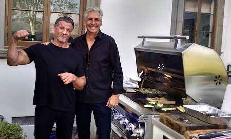 actor Sylvester Stallone flexing his muscle next to his big stainless steel gas grill