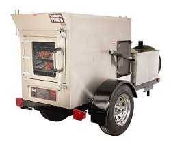 Large shiny steel box smoker on a trailer with big wheels. A door with window and large handle is on the back.
