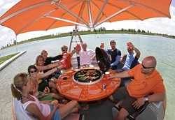 A group of people sitting in a circle in a round boat on a body of water. There is a round orange table in the middle of the boat with a black grill at the center. A large orange umbrella is overhead.