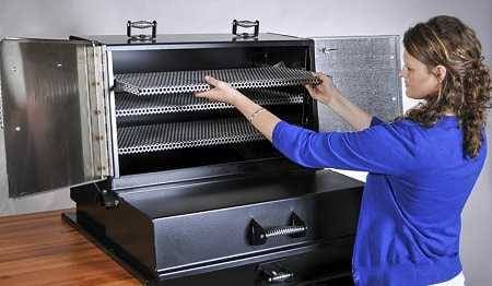 A woman in a blue shirt stands in front of a large black cabinet with double doors open. She is sliding a rack out of the top