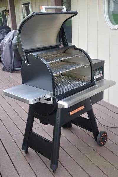 Large pellet smoker on an outdoor deck. The lid is up showing three slide out cooking grates. A wire plugged into a control panel on the right rests on the lower grate.