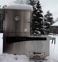 Large shiny smoker out side during the winter covered with snow. Snow covered pine trees are in the background.