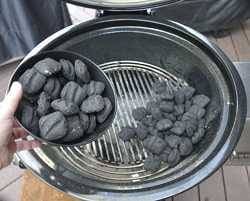 Black charcoal kettle grill with lid up and a pile of charcoal at the bottom. A hand on the left holds a black dish filled with more charcoal.