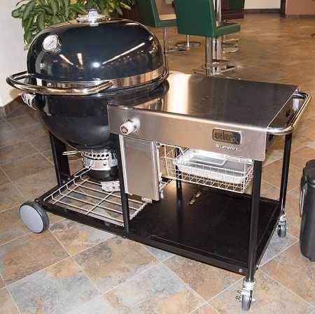 Large black charcoal kettle indoors on a showroom floor, mounted on a cart with four wheels.