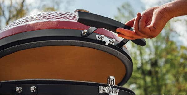 A hand effortlessly lifts the dome of a red kamado with fingertips.