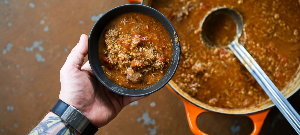 A bowl of Brunswick stew next to a simmering pot with a ladle.