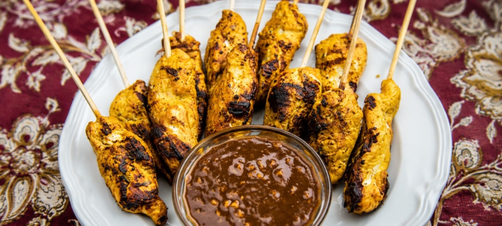 Chicken satay on a white plate along with a small dish of peanut sauce.