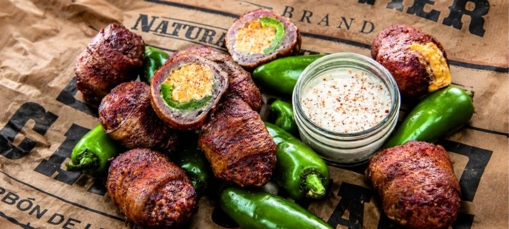 Armadillo egg of cheese and jalapeno stuffed bacon wrapped sausage and a bowl of sauce on newspaper.