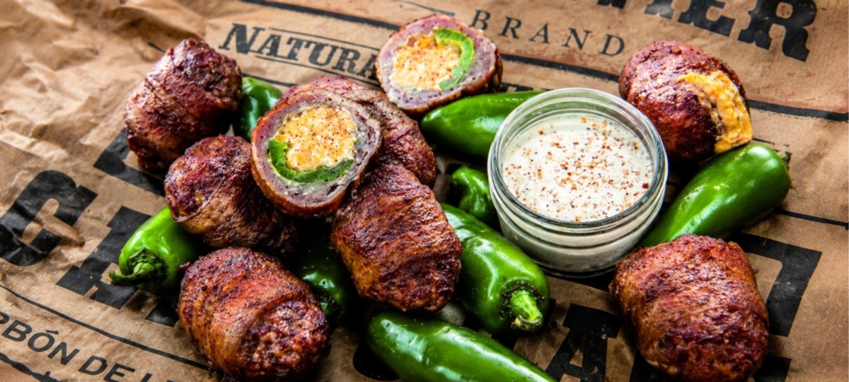 Armadillo egg of cheese and jalapeno stuffed bacon wrapped sausage and a bowl of sauce on newspaper