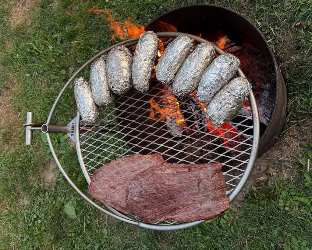 flank steak and foil wrapped potatoes on a grill grate over a camp fire