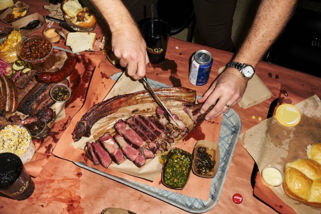 Smoked tomahawk steak on table with hands and knife cutting into steak.