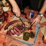 Smoked tomahawk steak on table with hands and knife cutting into steak.