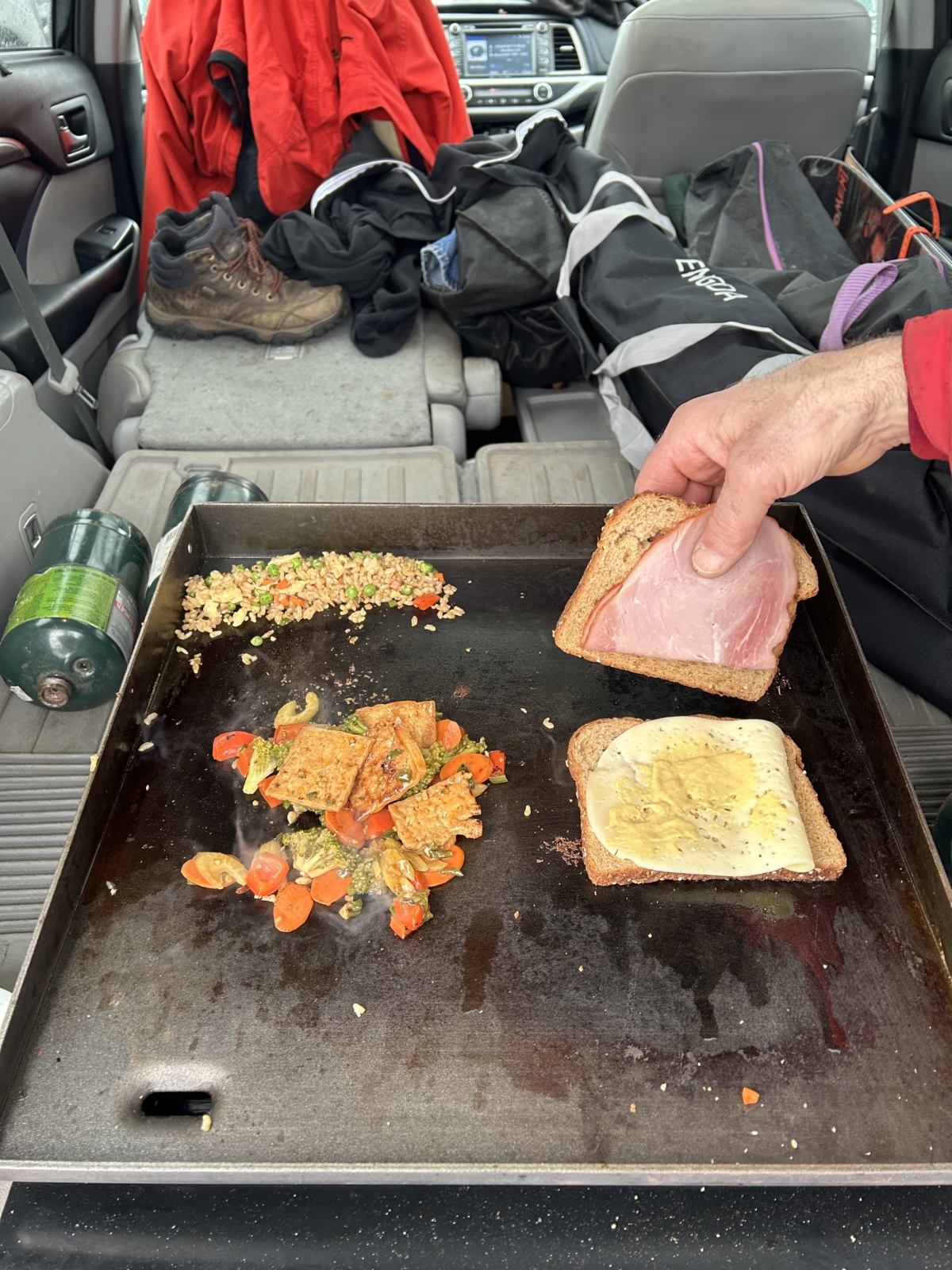 stir-fried vegetables and rice on a griddle with a man's hand making a ham and cheese sandwich alongside it, all in the back of a car.