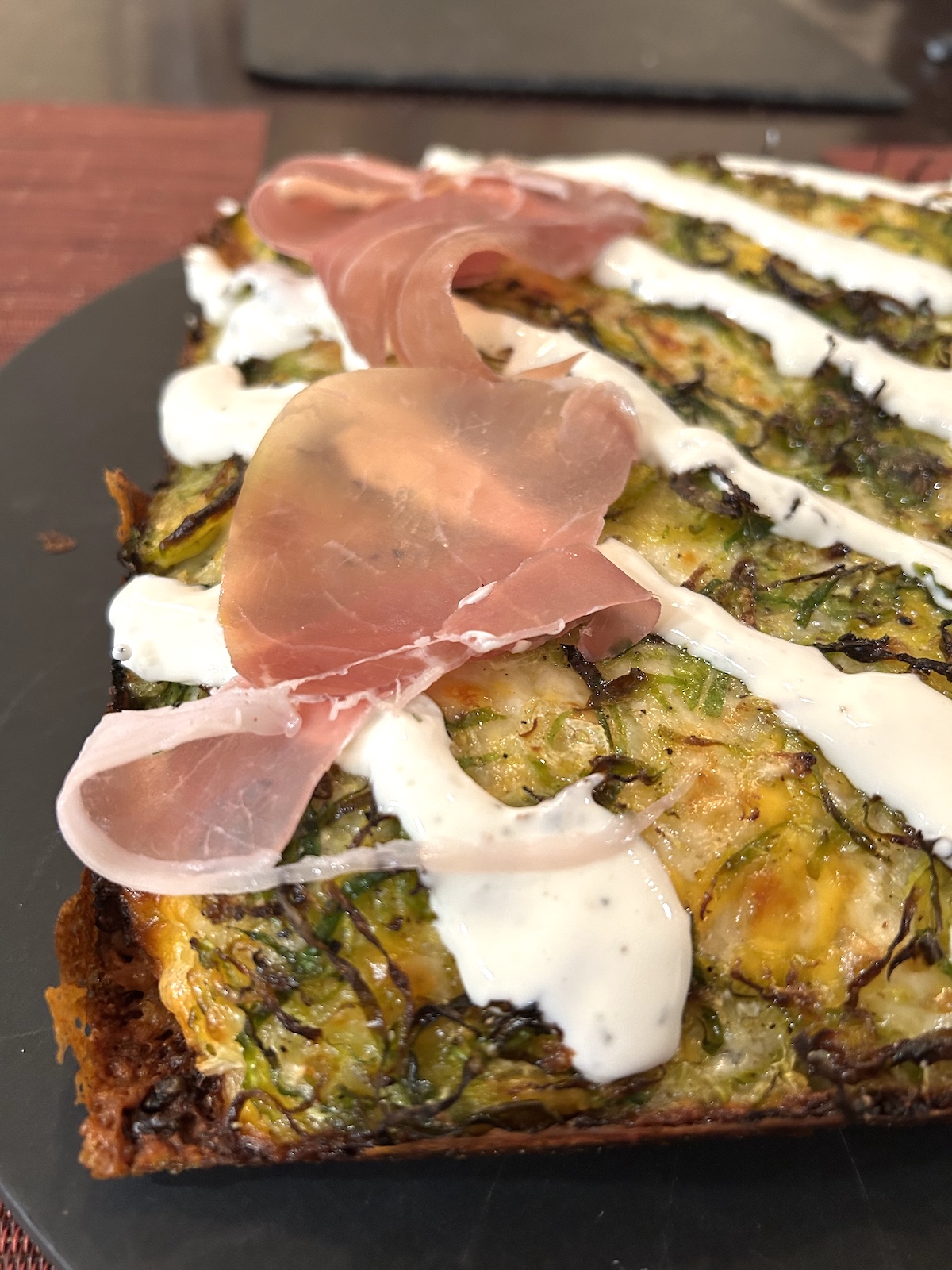 A closeup of a baked square Detroit style pizza topped with caramelized onions, shredded Brussels sprouts, Boursin cheese white sauce, and prosciutto.